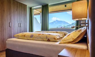 A cozy bedroom with a large window and a view of the mountains. There is a bed with colorful pillows and a warm atmosphere.