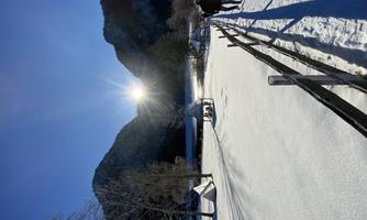 A winter landscape with a snow-covered ground and tall mountains in the background. The sun is shining brightly and a visitor is walking along a path beside a fence.
