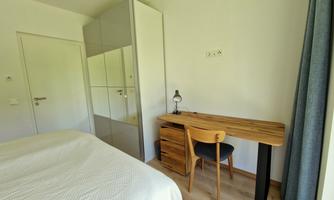 A bright room with a bed, a desk, and a wooden chair. There is a mirror on the wall and the windows are decorated with curtains.