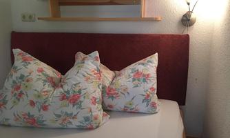 A cozy bed with large, blooming pillows. In the background, a simple shelf and a wall lamp can be seen.
