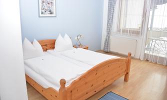 A bright bedroom with a wooden bed and two white duvets. A picture hangs on the wall, and large windows provide plenty of light.