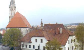 A historical cityscape with a church in the background and an old building in the foreground. Trees line the street, adding a pleasant atmosphere to the scene.