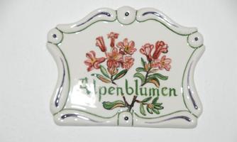 A decorative ceramic plaque with a floral illustration and the inscription "Alpenblumen". The colors are soft and harmonious.
