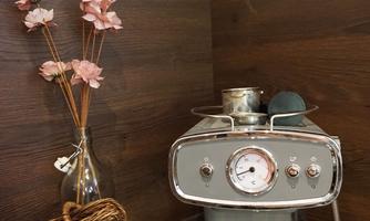 A modern espresso machine is sitting on a wooden surface. Next to it is a vase with pink flowers and a basket.