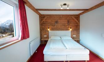 A cozy bedroom with a double bed and wooden-paneled walls. The room has large windows overlooking the mountains and a red carpeted floor.