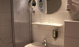 A modern bathroom with a shower, a sink, and two mirrors. The walls are bright tiled, and there is a small shelf with toiletries.