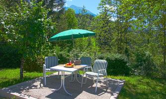 An inviting outdoor seating area with a table, chairs, and a sunshade. The surroundings are green and surrounded by trees, with a mountain landscape in the background.