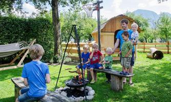 A group of children is sitting around a fire outdoors. In the background, a wooden structure and a hammock can be seen.