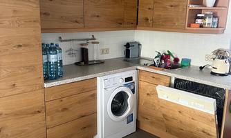 A modern kitchen with wooden furniture and a washing machine. Some kitchen utensils and a bottle of water are on the countertop.