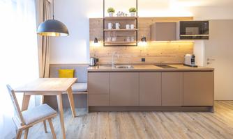 A modern kitchen with wooden shelves and a dining area. The room is bright and welcoming with a large window.