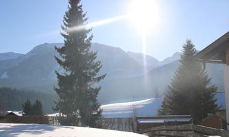 A wintry landscape with snow-covered mountains and trees. The sun shines brightly in the sky.