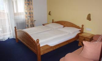 A cozy bedroom with a wooden double bed and a comfortable armchair. Large windows provide plenty of daylight.