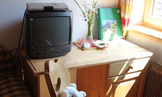 A small room with a wooden wardrobe and an old television. On the table are flowers and a few personal items.