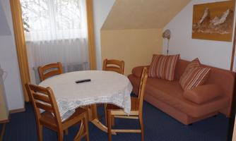 A cozy living room with a round table and a sofa. The room has large windows that let in plenty of light.