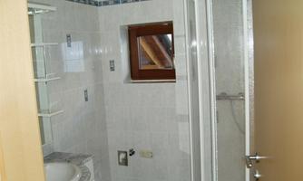 A bright bathroom with a shower, a sink, and a window. The walls are tiled in light colors and there are shelves for storage.