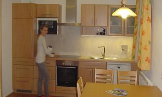 A modern kitchen with wooden furniture and a dining table. A person is standing at the sink and preparing something.