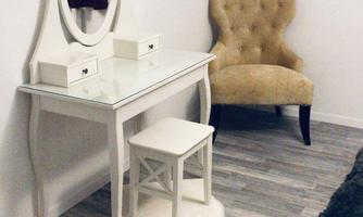 An elegant vanity table with a round mirror and two small drawers. Next to the table is a beige chair on a wooden floor.