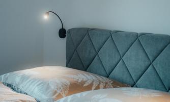 A modern bedroom with a stylish gray headboard and an art wall. The bedding is in soft tones and a wall lamp provides warm light.