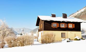 A charming house in winter, surrounded by freshly fallen snow. In the background, snow-covered mountains and a bright blue sky can be seen.