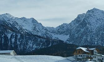 A wintry mountain landscape with snow-covered peaks. In the foreground, some houses can be seen on snow-covered ground.
