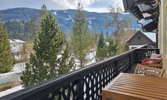 A balcony with wooden chairs overlooking a wintry landscape. In the background, green trees and snow-covered mountains can be seen.