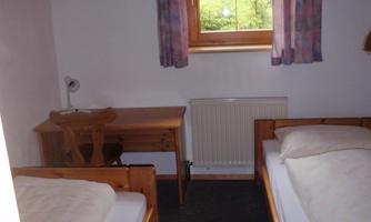 A simple bedroom with two single beds and a desk. It has a window with a view outside and curtains.
