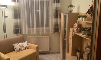 A cozy room with a beige sofa and a shelf cabinet. The windows are decorated with sheer curtains and there are some plants.