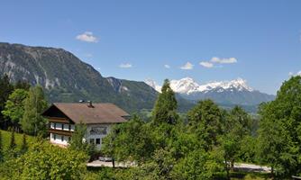 A picturesque landscape with a traditional house surrounded by trees. In the background, majestic mountains with snow-capped peaks are visible under a blue sky.