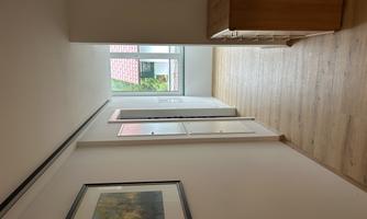 A bright hallway with wooden floors and large windows. Pictures hang on the walls, and a wooden cabinet is on the right side.