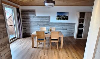 A bright dining room with a wooden table and four chairs. The walls are clad with wooden panels and there are large windows that let in plenty of daylight.