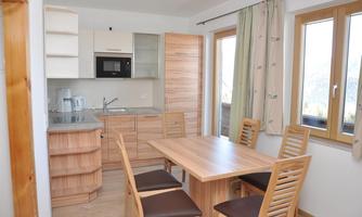 A modern kitchen with wooden shelves and a dining area. The room is bright and friendly with large windows.