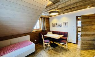 A cozy room with wooden paneling and a sloped ceiling. There is a bed, a dining table with chairs, and large windows that let in natural light.