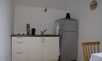A small kitchen with a refrigerator and a sink. On the left, there is a table with a chair.