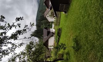 A green meadow with several houses in the background. The sky is cloudy and a hilly landscape surrounds it.