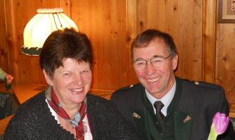 A smiling couple sits in a cozy room with wooden paneling. In the background, a lamp can be seen that creates a warm atmosphere.