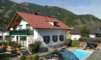 A beautiful house with a red roof, surrounded by mountains. In the foreground, there is a pool and well-maintained gardens.