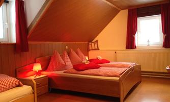 A cozy bedroom with wood paneling and red curtains. There are two beds with checked pillows and a nightstand with a lamp.