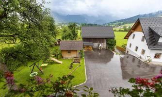 An idyllic farmhouse surrounded by lush greenery and mountains in the background. The yard is well-maintained and a playground for children is visible.
