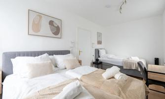 A modern bedroom with two beds, a large window front, and decorative cushions. The walls are painted in soft colors and there are artworks for decoration.