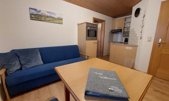 A cozy living room with a blue sofa and a wooden table. In the corner, there is a modern kitchenette.