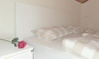 A simple bedroom with a bed and pillows in neutral colors. On the nightstand lies a red rose.