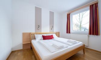 A bright bedroom with a double bed and decorative cushions. The large window lets in daylight and offers a view of the trees outside.