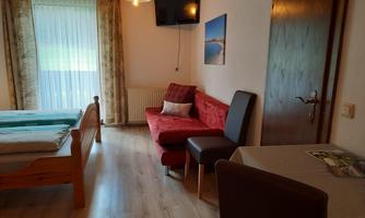 A cozy room with two beds, a red sofa, and a small table. The window lets in plenty of daylight and there is a television on the wall.