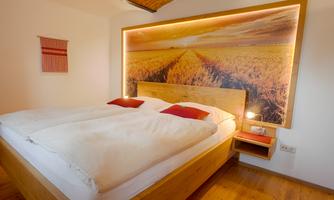 A cozy bedroom with a large double bed and red pillows. The wall behind the bed features a picture of a landscape painting with wheat fields.