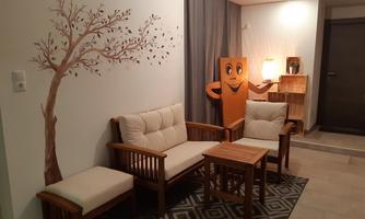 A cozy living area with wooden furniture and a bright carpet. A decorative tree is visible on the wall.