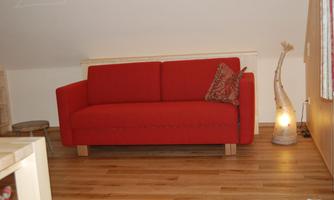 A red sofa is in a cozy room with wooden flooring. A decorative lamp and a small table complete the decor.