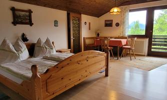 A cozy room with a wooden bed and a dining area. Large windows provide a view of the green landscape.