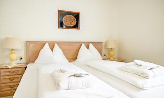 A bright room with two single beds, clean bedding, and bedside lamps. A small painting hangs on the wall.
