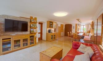 A bright living room with a red sofa and a wooden TV unit. The room has a dining area with a large table and windows that let in plenty of light.