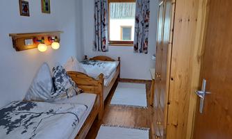 A simple bedroom with two wooden single beds. The walls are light and there is a window with curtains.